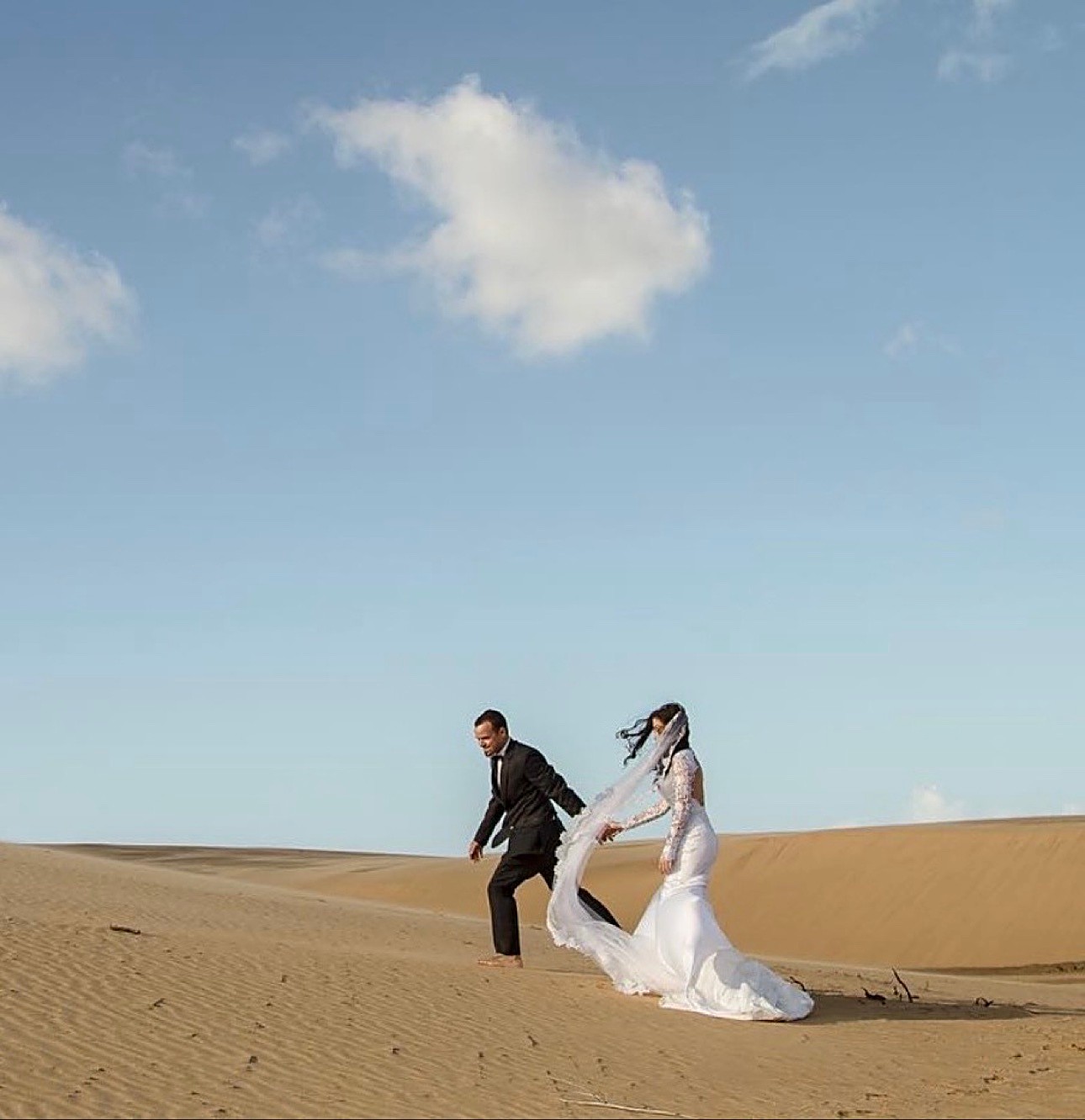 Newlyweds walking barefoot together across sweeping sand dunes