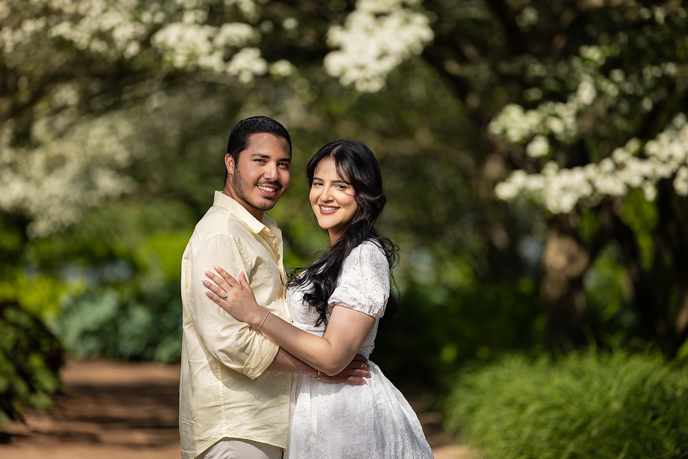 Engaged couple embracing beneath flowering trees in a sunlit garden, her new engagement ring resting on his shoulder — engagement session portrait by Abigail Salazar