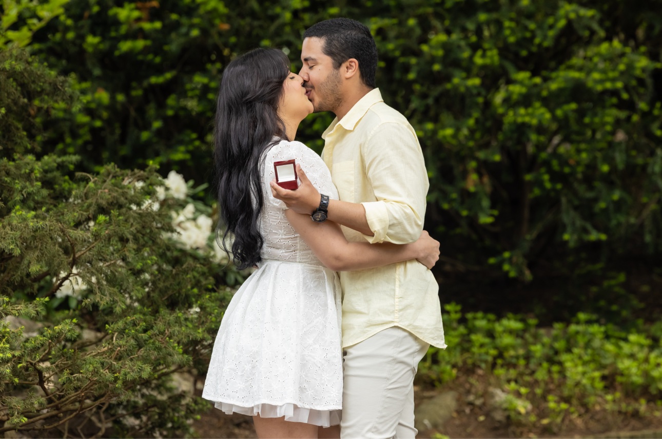 The newly engaged couple kissing among green garden shrubs, the groom holding the red ring box in his hand