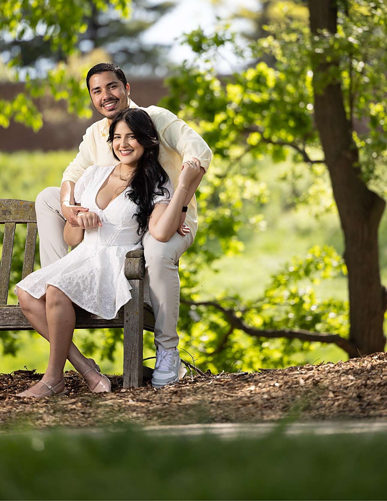 The couple seated on a wooden garden bench in golden light, the groom behind her with his arms around her, both smiling